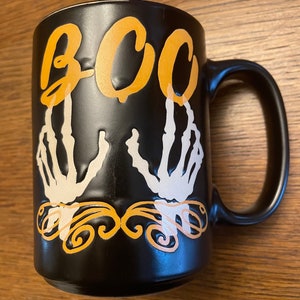 Puede incluir: Taza de caf&eacute; de cer&aacute;mica negra con un dise&ntilde;o de mano de esqueleto blanco y la palabra "BOO" en letras naranjas.