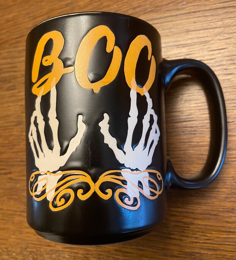 Puede incluir: Taza de caf&eacute; de cer&aacute;mica negra con un dise&ntilde;o de mano de esqueleto blanco y la palabra "BOO" en letras naranjas.