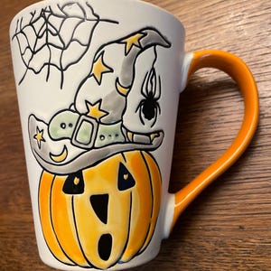 Puede incluir: Una taza de cer&aacute;mica blanca con asa naranja presenta un dise&ntilde;o pintado a mano de una calabaza con un sombrero de bruja con estrellas y una ara&ntilde;a.