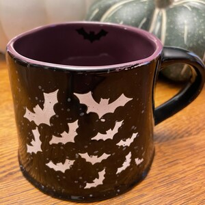 Puede incluir: Taza de cer&aacute;mica negra con un borde morado y un dise&ntilde;o de murci&eacute;lago blanco. La taza est&aacute; decorada con murci&eacute;lagos blancos sobre un fondo negro.