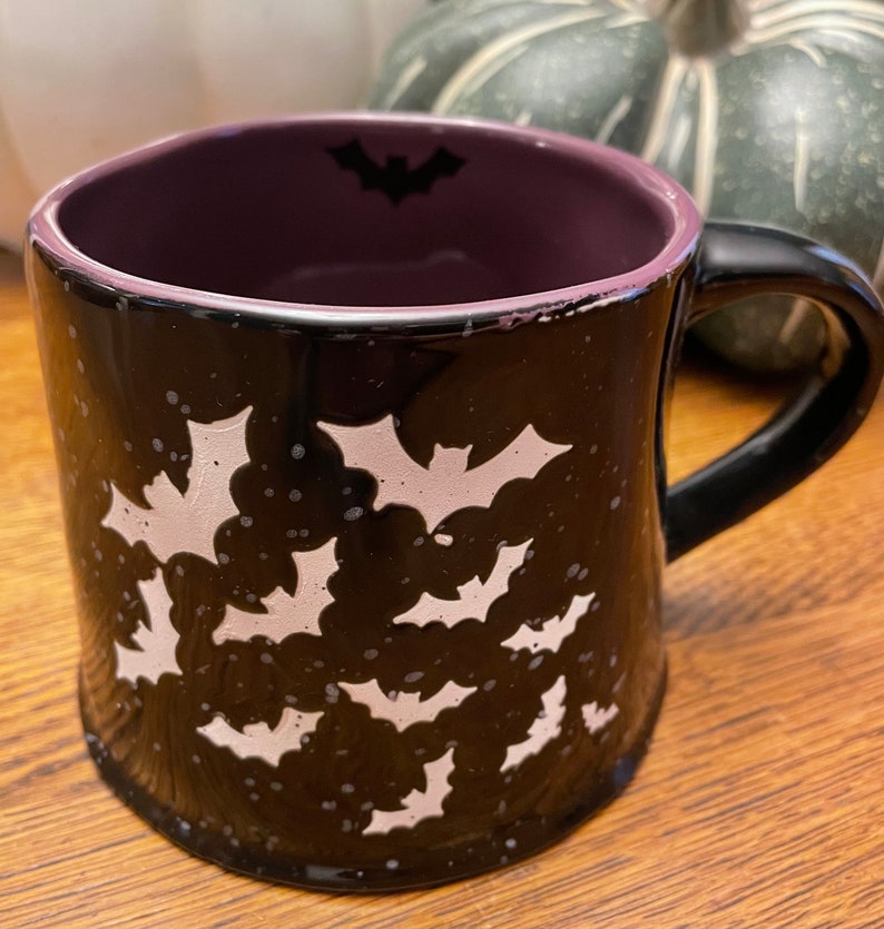 Puede incluir: Taza de cer&aacute;mica negra con un borde morado y un dise&ntilde;o de murci&eacute;lago blanco. La taza est&aacute; decorada con murci&eacute;lagos blancos sobre un fondo negro.