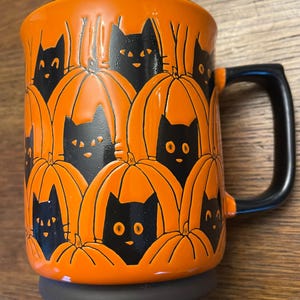 Puede incluir: Taza de cer&aacute;mica naranja con caras de gatos negros asomando desde detr&aacute;s de calabazas naranjas. La taza tiene un asa negra.