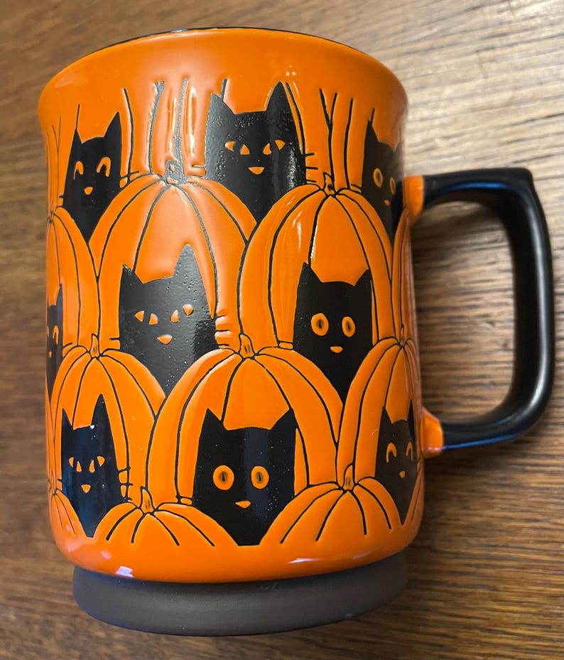 Puede incluir: Taza de cer&aacute;mica naranja con caras de gatos negros asomando desde detr&aacute;s de calabazas naranjas. La taza tiene un asa negra.