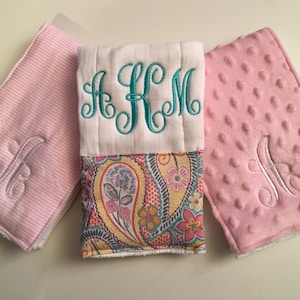 May include: Three personalized baby items: a white and paisley burp cloth with teal monogram, and two pink items with the letter "A" embroidered. One pink item has a striped pattern, the other has a dimpled texture.