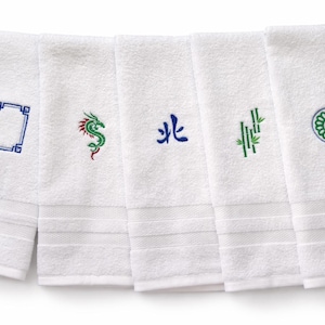 May include: Five white rectangular hand towels arranged in a row, each featuring a distinct embroidered design. The motifs include a blue geometric frame, a green and red dragon, a blue Chinese character, green bamboo stalks, and a circular floral emblem.