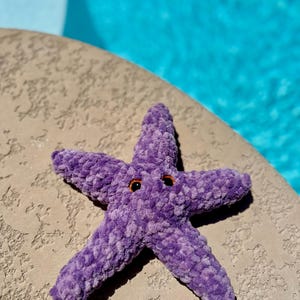 May include: A plush purple starfish with black eyes. The starfish is lying on a light brown textured surface.