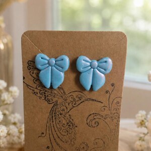 May include: A pair of light blue bow-shaped stud earrings with a subtle pearlescent finish. The earrings are attached to a rectangular brown card featuring a black line-art illustration of a hummingbird and floral flourishes.