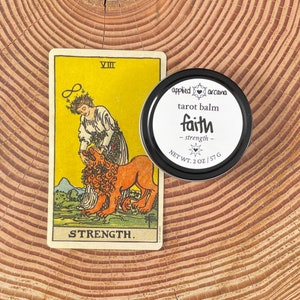 May include: A Strength tarot card featuring a figure with a lion and a round black tin of Applied Arcana tarot balm labeled faith strength with a net weight of 2 oz. The card displays the Roman numeral VIII and the word STRENGTH.