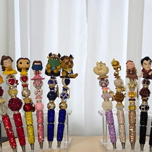 May include: A collection of decorative pens featuring character figurines. The pens have jewel-encrusted barrels in various colors, including red, blue, and gold. The figurines include characters from a popular animated film, adding a whimsical touch to the writing instruments.