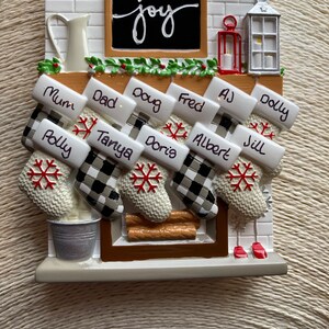 Puede incluir: Un adorno de chimenea de cerámica blanca con una pizarra que dice "joy" y calcetines con nombres escritos en ellos. Los calcetines son blancos con patrones de cuadros negros y blancos y copos de nieve rojos.