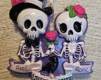 May include: A decorative ornament featuring two skeleton figures, one wearing a top hat and the other a floral crown. They hold a rose, with a black crow and the text "LOVE YOU TO DEATH" on a banner. The names "Axel" and "Rose" are also present.