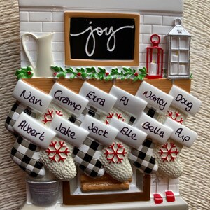 Puede incluir: Un adorno de chimenea de cerámica blanca con una pizarra negra que dice "joy" y ocho calcetines con nombres escritos en ellos: Nan, Gramp, Ezra, PJ, Wancy, Doug, Albert, Jake, Jack, Elle, Bella, Pup.