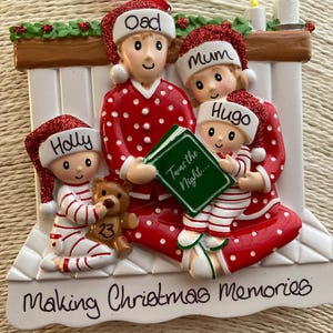 May include: A festive Christmas ornament depicting a family of four in red and white pyjamas and Santa hats. The ornament reads "Making Christmas Memories" and includes the names "Dad", "Mum", "Holly", and "Hugo".