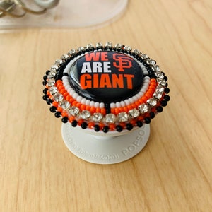May include: A black and orange phone grip with a white and black button. The grip is decorated with rhinestones and beads. The button has a black background with white text that reads "WE ARE GIANT" and a red "SF" logo.