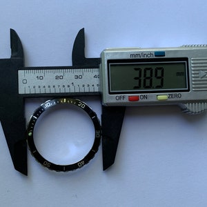 Może przedstawiać: Czarny bezel z białymi oznaczeniami dla tarczy zegarka, zmierzony cyfrowym suwmiarką, pokazującym pomiar 38,9 mm.