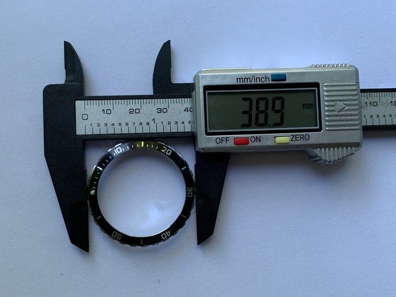 Może przedstawiać: Czarny bezel z białymi oznaczeniami dla tarczy zegarka, zmierzony cyfrowym suwmiarką, pokazującym pomiar 38,9 mm.