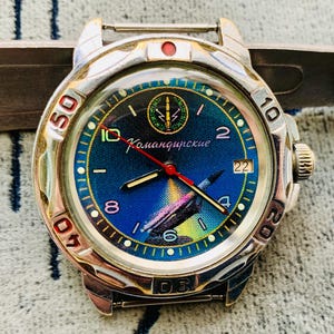 Op de afbeelding: Een zilverkleurig polshorloge met een blauwe wijzerplaat met een raketillustratie en de Cyrillische tekst "Командирские". Het horloge heeft een draaibare ring met cijfers en een rode secondewijzer. Vintage stijl.