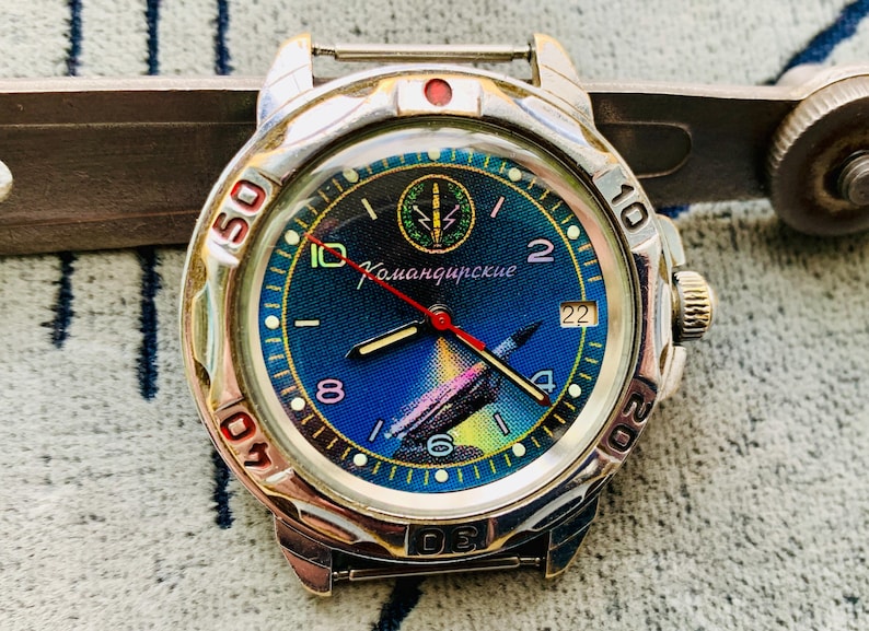 Op de afbeelding: Een zilverkleurig polshorloge met een blauwe wijzerplaat met een raketillustratie en de Cyrillische tekst "Командирские". Het horloge heeft een draaibare ring met cijfers en een rode secondewijzer. Vintage stijl.