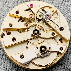 K&ouml;nnte beinhalten: Nahaufnahme eines Vintage-Uhrwerks mit komplizierten Zahnr&auml;dern und Komponenten. Der Mechanismus verf&uuml;gt &uuml;ber goldfarbene Platten, sichtbare Schrauben und den Text "Twenty 20 Jewels". Das Uhrwerk ist eine komplexe Anordnung von Pr&auml;zisionsteilen.