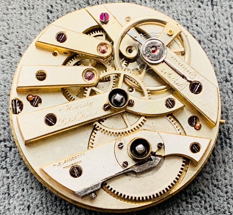 K&ouml;nnte beinhalten: Nahaufnahme eines Vintage-Uhrwerks mit komplizierten Zahnr&auml;dern und Komponenten. Der Mechanismus verf&uuml;gt &uuml;ber goldfarbene Platten, sichtbare Schrauben und den Text "Twenty 20 Jewels". Das Uhrwerk ist eine komplexe Anordnung von Pr&auml;zisionsteilen.