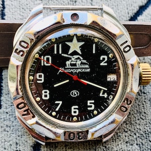 May include: Close-up of a vintage wristwatch with a black face and silver bezel. The watch face features white numerals, a star, and a tank graphic. The hands are red and white. The watch has a gold-colored crown and a brown strap.