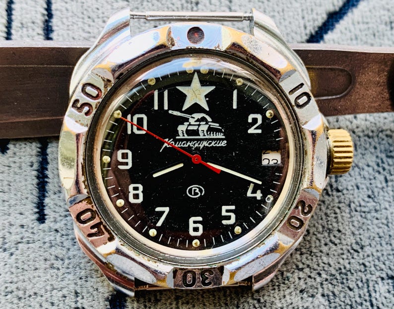 May include: Close-up of a vintage wristwatch with a black face and silver bezel. The watch face features white numerals, a star, and a tank graphic. The hands are red and white. The watch has a gold-colored crown and a brown strap.