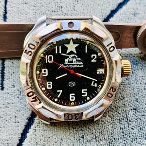 May include: A vintage wristwatch with a black face, white numerals, and a red second hand. The watch has a silver-colored bezel with a rotating outer ring. The face features a star and a tank graphic. The watch is a military-style timepiece.