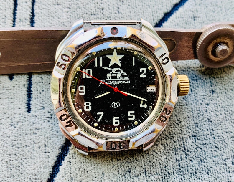 May include: A vintage wristwatch with a black face, white numerals, and a red second hand. The watch has a silver-colored bezel with a rotating outer ring. The face features a star and a tank graphic. The watch is a military-style timepiece.