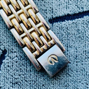 May include: Close-up of a two-tone watch band with gold and silver links. The watch band is made of metal and has a clasp with a logo. The watch band is on a blue textured surface.