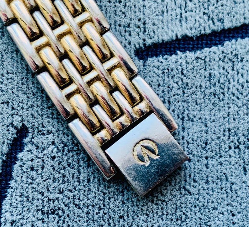 May include: Close-up of a two-tone watch band with gold and silver links. The watch band is made of metal and has a clasp with a logo. The watch band is on a blue textured surface.