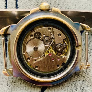 Op de afbeelding: Close-up van de open achterkant van een vintage polshorloge, met ingewikkelde mechanische tandwielen en componenten. De horlogekast heeft een goudkleurige afwerking met tekenen van slijtage. De interne mechanismen omvatten kleine, gedetailleerde tandwielen en rode juwelen.