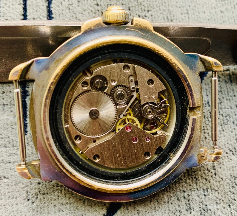 Op de afbeelding: Close-up van de open achterkant van een vintage polshorloge, met ingewikkelde mechanische tandwielen en componenten. De horlogekast heeft een goudkleurige afwerking met tekenen van slijtage. De interne mechanismen omvatten kleine, gedetailleerde tandwielen en rode juwelen.