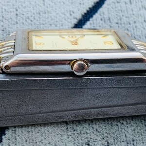 May include: A rectangular wristwatch with a silver-colored case and a gold-colored band. The watch face is cream-colored with black numerals. The watch is resting on a dark gray surface.