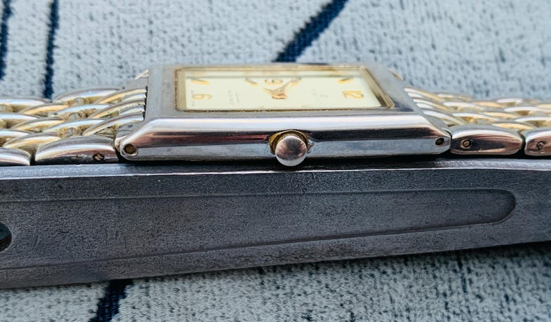 May include: A rectangular wristwatch with a silver-colored case and a gold-colored band. The watch face is cream-colored with black numerals. The watch is resting on a dark gray surface.