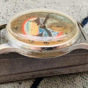 Puede incluir: Reloj de pulsera vintage con caja plateada y cristal abovedado transparente. La esfera del reloj presenta un dise&ntilde;o de paisaje colorido con una peque&ntilde;a subesfera. Las manecillas son negras y el reloj est&aacute; sujeto a una correa plateada.