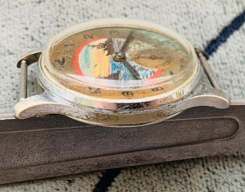 Puede incluir: Reloj de pulsera vintage con caja plateada y cristal abovedado transparente. La esfera del reloj presenta un dise&ntilde;o de paisaje colorido con una peque&ntilde;a subesfera. Las manecillas son negras y el reloj est&aacute; sujeto a una correa plateada.