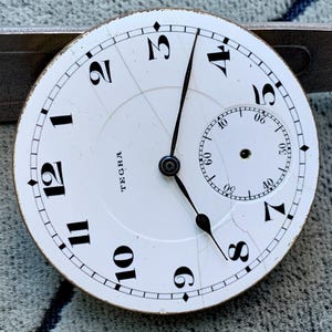 Puede incluir: Esfera de reloj blanca antigua con n&uacute;meros y manecillas negras. La esfera tiene una subesfera m&aacute;s peque&ntilde;a para los segundos y la palabra "TEGRA" impresa. La esfera est&aacute; agrietada y muestra signos de edad.