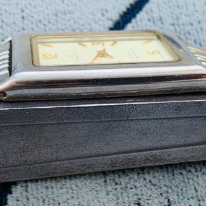 May include: A rectangular wristwatch with a silver-colored case and a gold-colored band. The watch face is cream-colored with gold-colored hands and numerals. The watch is resting on a dark gray surface.