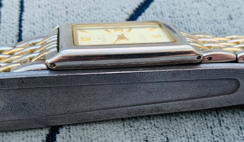 May include: A rectangular wristwatch with a silver-colored case and a gold-colored band. The watch face is cream-colored with gold-colored hands and numerals. The watch is resting on a dark gray surface.