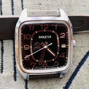 Puede incluir: Un reloj de pulsera cuadrado plateado con una esfera negra. La esfera del reloj tiene números blancos y un símbolo de martillo y hoz rojo. La esfera del reloj también tiene la palabra "PAKETA" impresa.