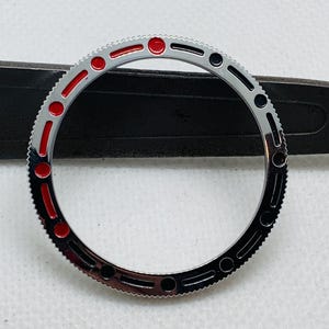 Op de afbeelding: Een zilveren horloge bezel met zwarte en rode markeringen. De bezel heeft een getextureerde rand en een glad, gepolijst oppervlak.