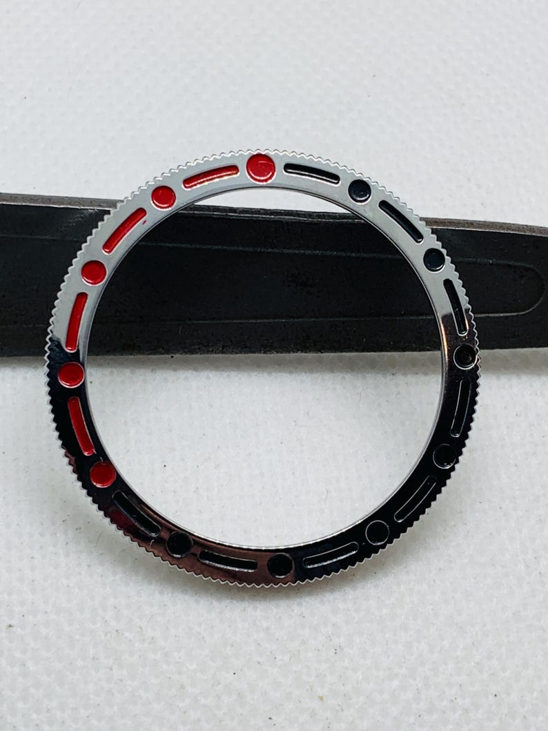 Op de afbeelding: Een zilveren horloge bezel met zwarte en rode markeringen. De bezel heeft een getextureerde rand en een glad, gepolijst oppervlak.