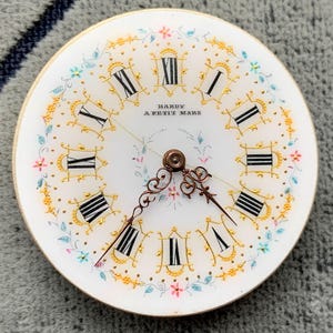 K&ouml;nnte beinhalten: Ein Vintage-Zifferblatt mit wei&szlig;em Hintergrund, schwarzen r&ouml;mischen Ziffern und aufwendigen goldenen Details. Das Zifferblatt hat zarte florale Akzente in Rosa, Blau und Gelb, mit der Aufschrift "HARDY A PETIT MARS". Die Zeiger sind dunkelbronzen.