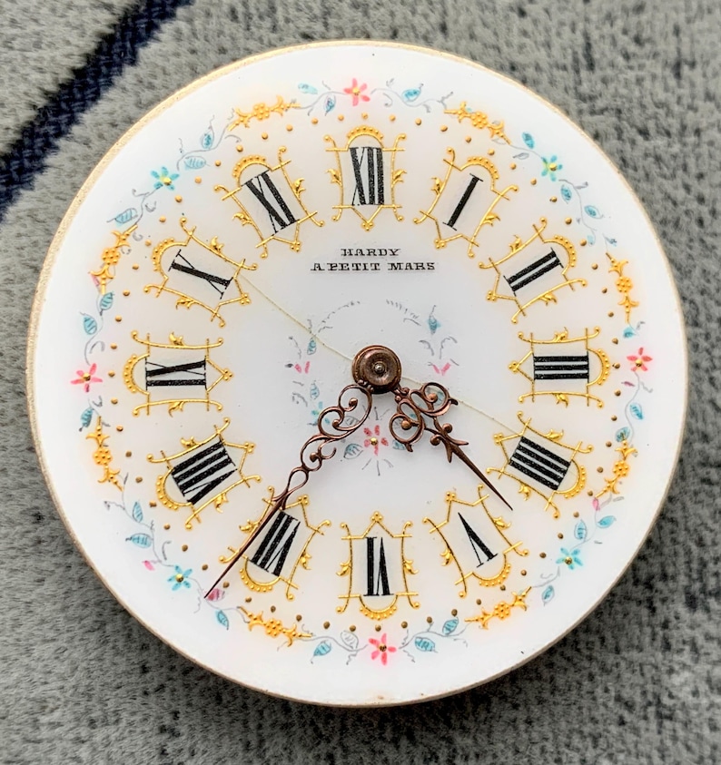 K&ouml;nnte beinhalten: Ein Vintage-Zifferblatt mit wei&szlig;em Hintergrund, schwarzen r&ouml;mischen Ziffern und aufwendigen goldenen Details. Das Zifferblatt hat zarte florale Akzente in Rosa, Blau und Gelb, mit der Aufschrift "HARDY A PETIT MARS". Die Zeiger sind dunkelbronzen.