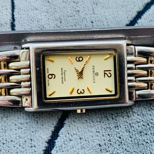 May include: A rectangular Perucci watch with a silver and gold band. The watch face is cream-colored with gold-tone hands and markers. The watch displays the numbers 3, 6, 9, and 12. The watch is marked "Expression" and "Water Proof".