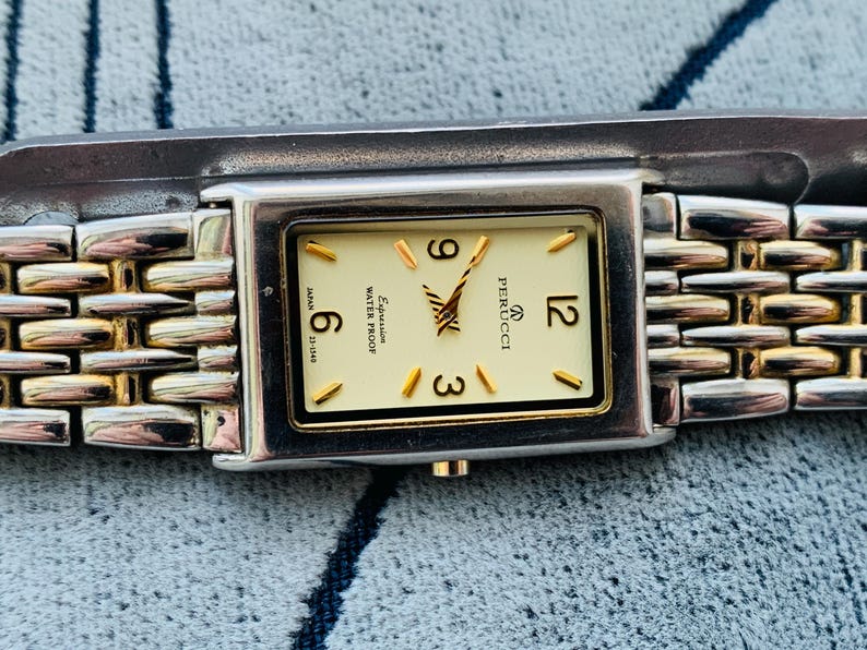 May include: A rectangular Perucci watch with a silver and gold band. The watch face is cream-colored with gold-tone hands and markers. The watch displays the numbers 3, 6, 9, and 12. The watch is marked "Expression" and "Water Proof".