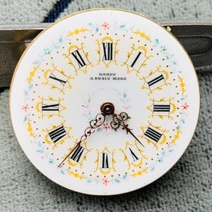 K&ouml;nnte beinhalten: Ein Vintage-Zifferblatt mit wei&szlig;em Hintergrund, schwarzen r&ouml;mischen Ziffern und zarten floralen Akzenten in Gold, Rosa und Blau. Die Worte "HARDY A PETIT MARS" sind in Schwarz gedruckt. Die Zeiger sind dunkelbronze.