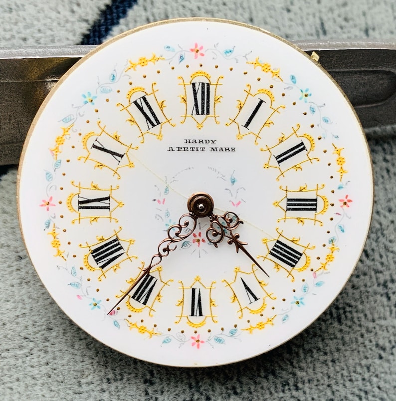 K&ouml;nnte beinhalten: Ein Vintage-Zifferblatt mit wei&szlig;em Hintergrund, schwarzen r&ouml;mischen Ziffern und zarten floralen Akzenten in Gold, Rosa und Blau. Die Worte "HARDY A PETIT MARS" sind in Schwarz gedruckt. Die Zeiger sind dunkelbronze.