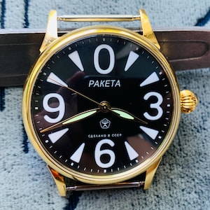 Puede incluir: Un reloj de pulsera dorado con esfera negra con números y manecillas blancas. La esfera del reloj muestra la palabra "RAKETA" y el texto "СДЕЛАНО В СССР". El reloj tiene una corona dorada y una fijación de correa metálica.
