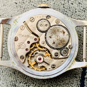 Puede incluir: Primer plano del mecanismo de un reloj vintage. La caja plateada enmarca los intrincados engranajes, joyas y muelles. La esfera del reloj muestra el texto "15 KAMHEЙ" y "423". El reloj tiene una peque&ntilde;a corona.